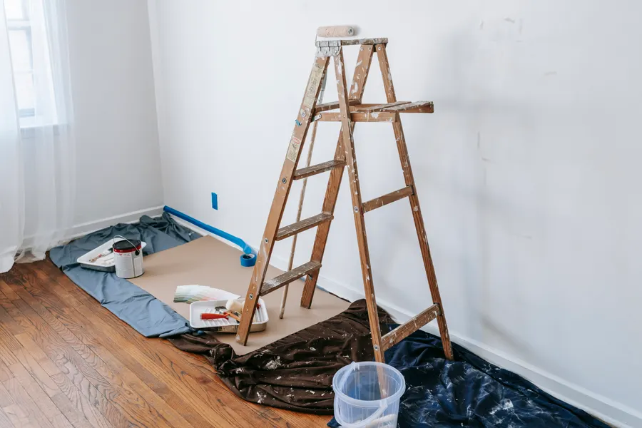 Unfurnished freshly painted room with installed baseboards, hardwood flooring covered by paper and drop cloths, and a ladder