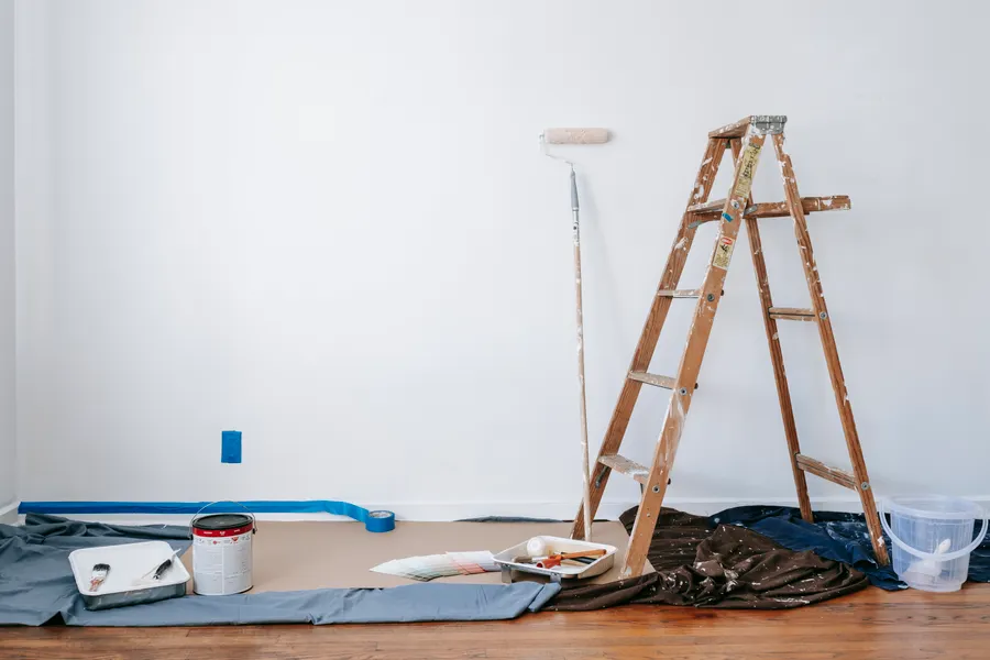 Unfurnished white room with a ladder, protected hardwood flooring, painting materials and blue painter’s tape