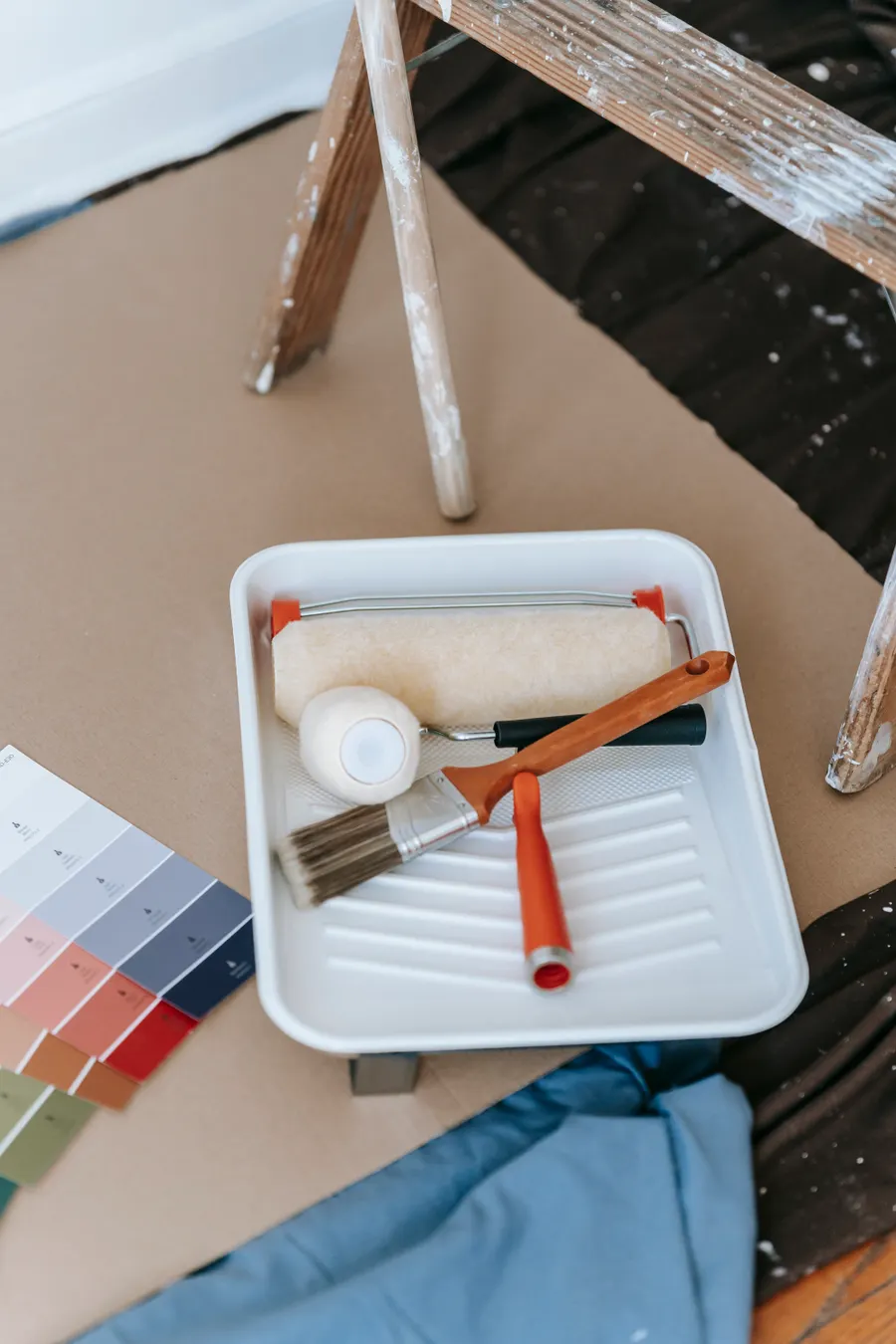 Paint tray, brushes and roller beside a ladder on floor protection paper and drop cloths, with no people or furniture